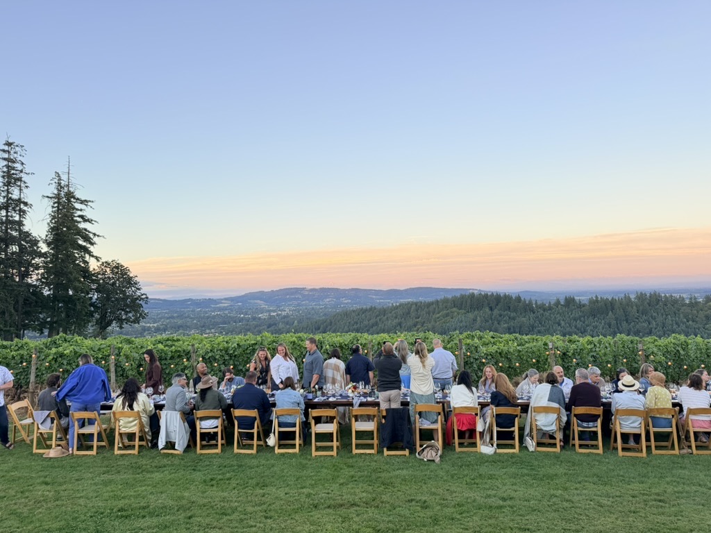 A ticketed dinner at sunset: guests seated along one long table at the edge of the vineyard, hills in the distance